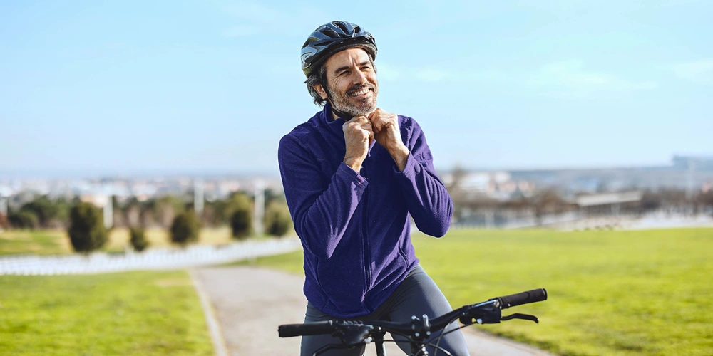 Lachender Radfahrer mit Helm auf einer Wiese, im Hintergrund eine Stadtlandschaft.